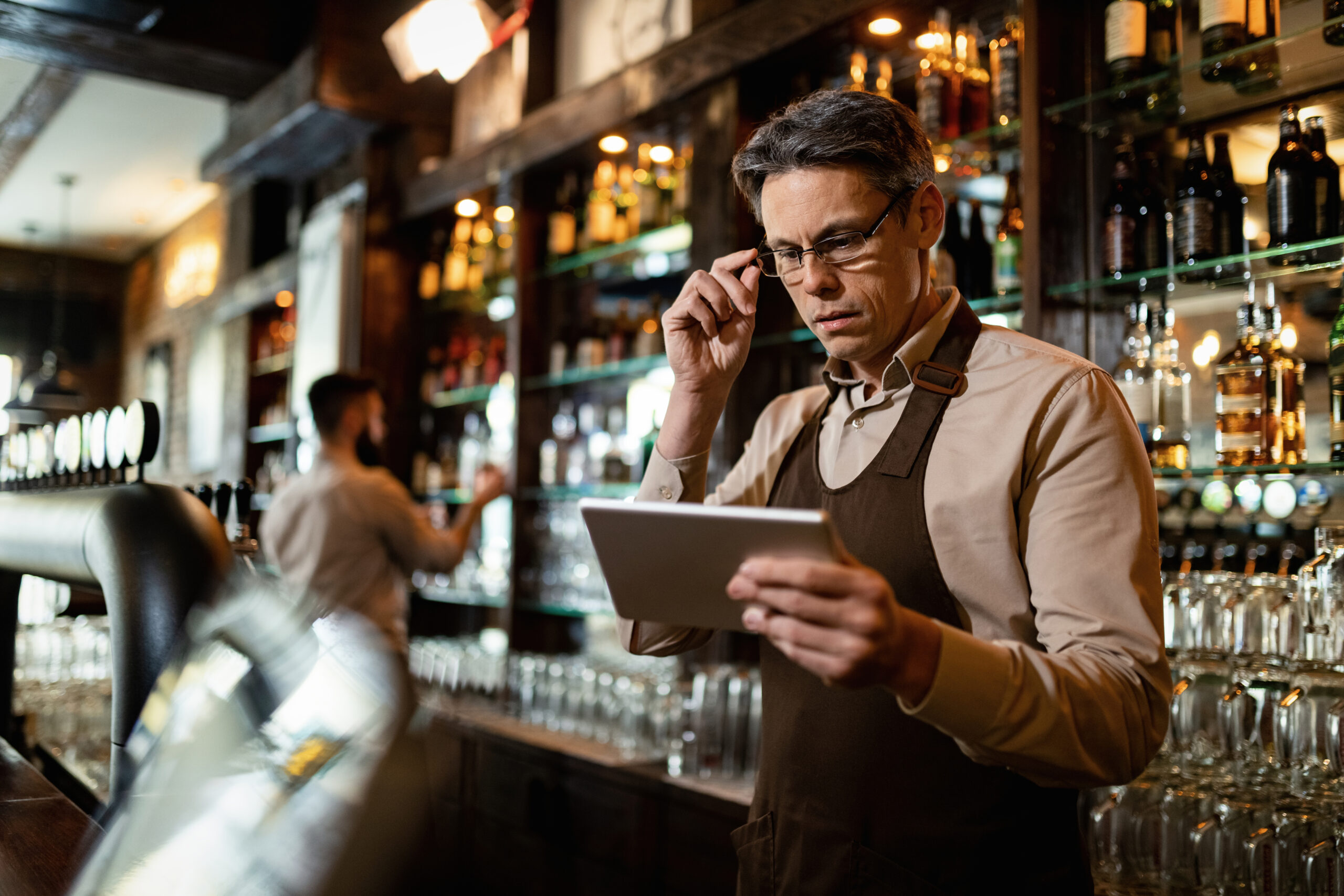A mid-adult bar manager wearing an apron and glasses reviews cost data on a digital tablet while standing in a bar. Shelves with bottles of alcohol and a bartender working in the background emphasize the hospitality setting. This image relates to managing theoretical COGS in hospitality to improve cost efficiency and profitability.